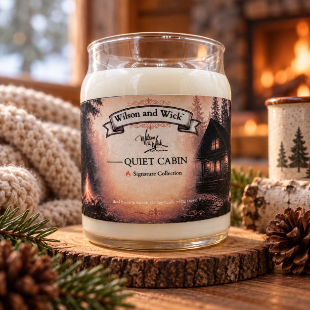 A glass jar candle labeled 'Wilson and Wick Quiet Cabin' sits on a rustic wood slice surrounded by pinecones, evergreen branches, and a cozy fireplace background.