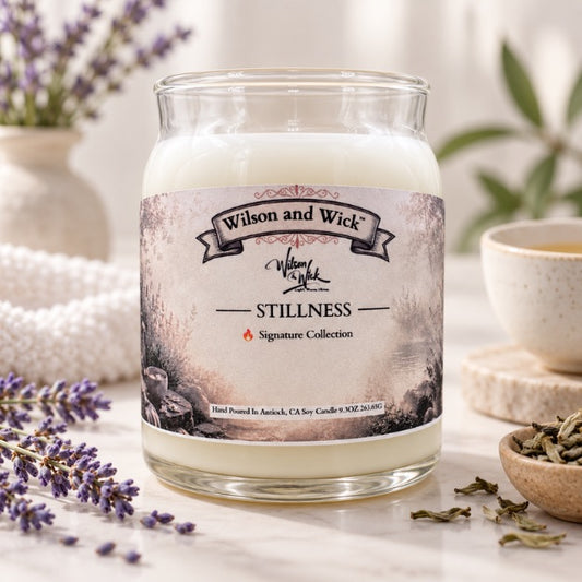 A glass jar candle labeled 'Wilson and Wick' and 'Stillness' sits on a light surface with lavender sprigs, a cup, and dried herbs in a softly styled setting.
